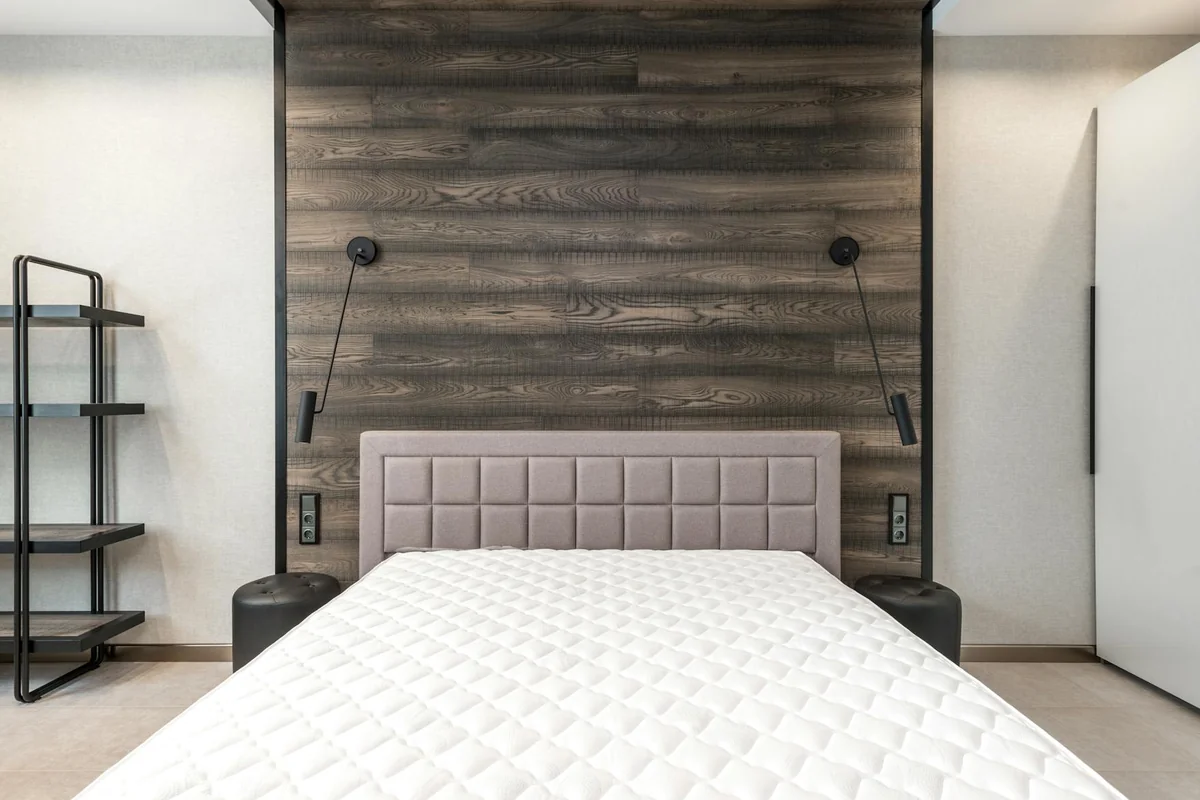 Comfortable bed with white mattress placed at wall with wooden element in stylish spacious bedroom with lamps and shelves in 