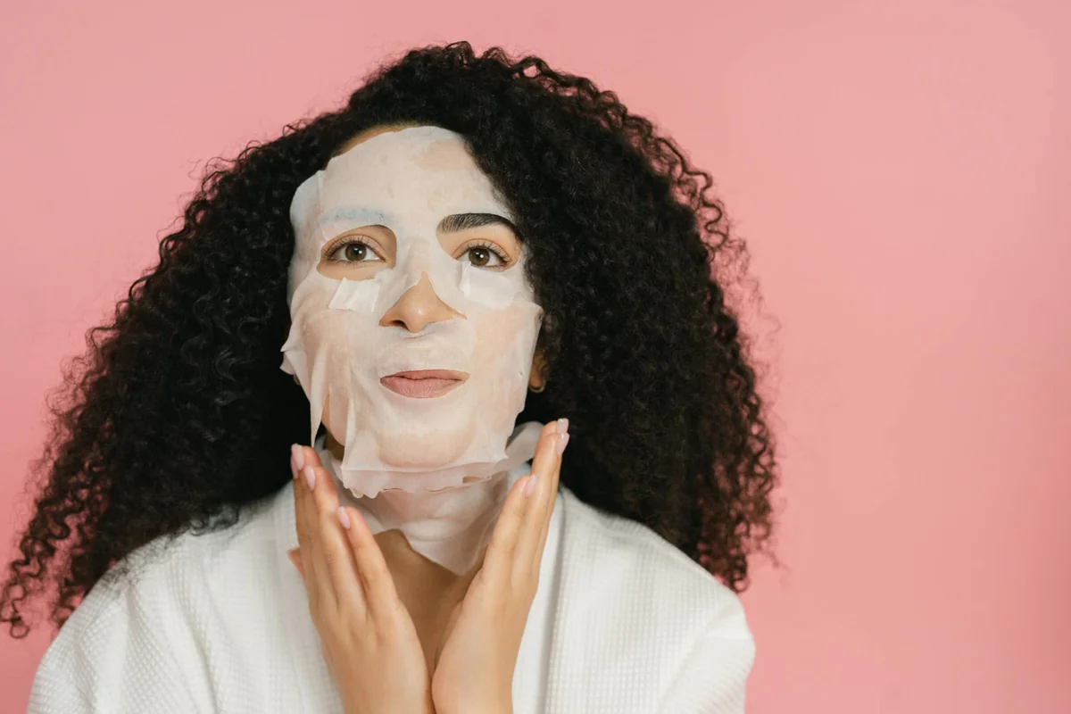 A woman in a bathrobe applying a moisturizing face mask on a pink background.