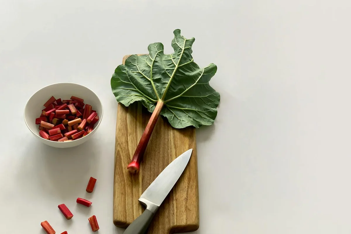 Fresh rhubarb stalk and leaves on a wooden cutting board with a knife, ready for cooking.