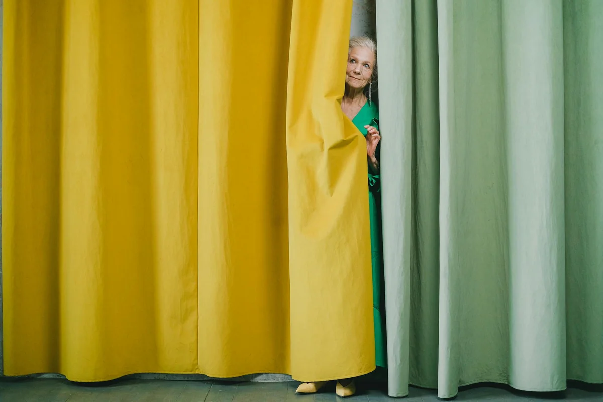 Elderly woman peeks from behind colorful curtains, creating a playful and vibrant scene.