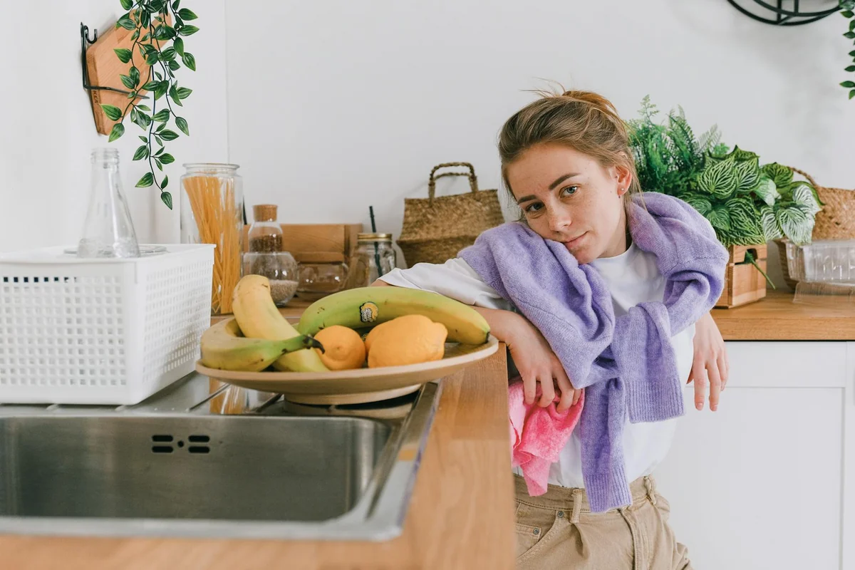 Positive young woman in casual clothes leaning on wooden counter near plate with fresh fruits and tableware in light kitchen 