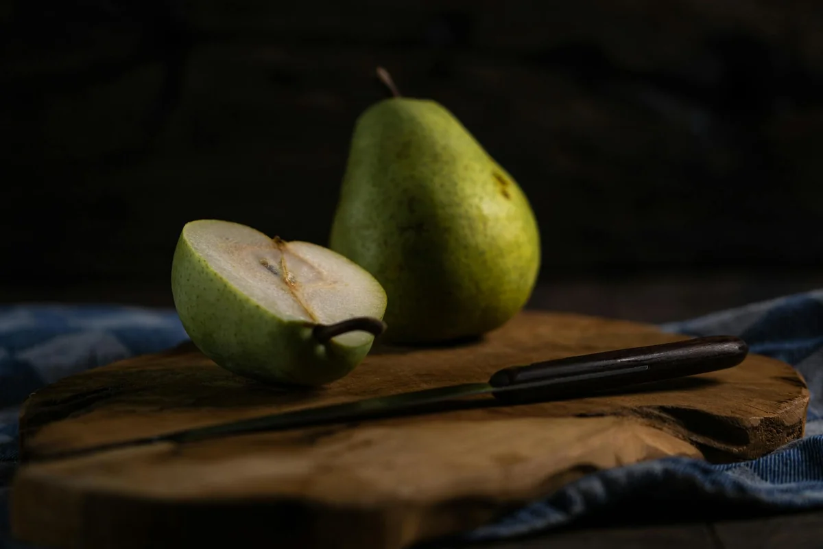 Fresh pears on a wooden board, one sliced with a knife, in a rustic setting.