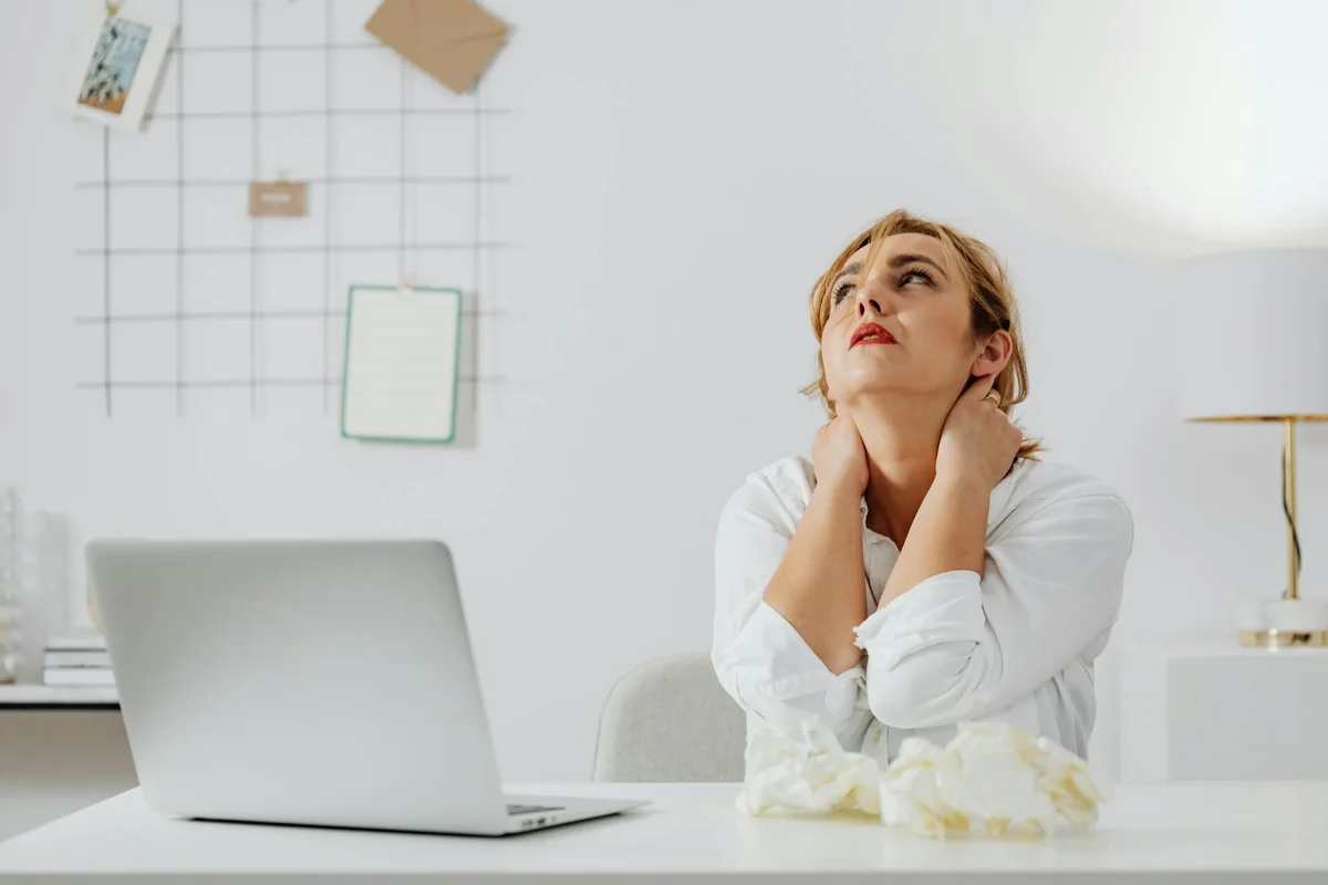A stressed woman in white long sleeves sits at a desk with a laptop, looking upwards in an office.