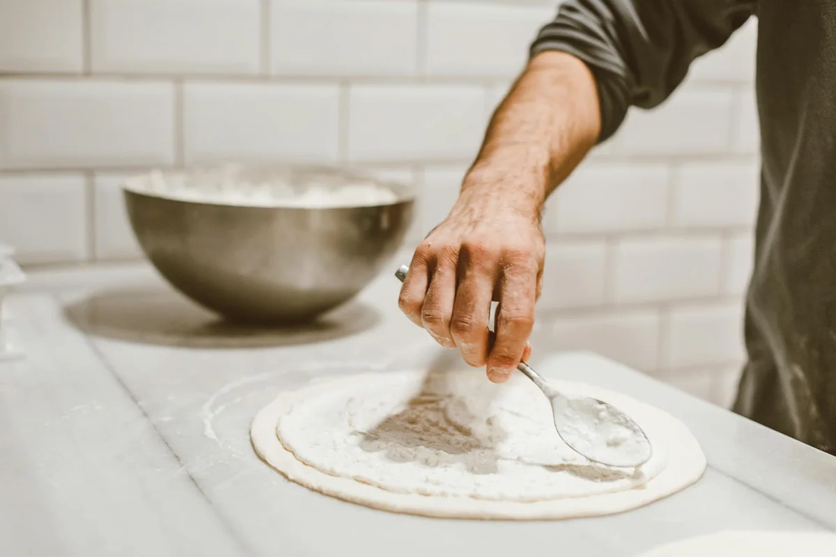A chef spreads white sauce on pizza dough with a spoon in a kitchen setting.