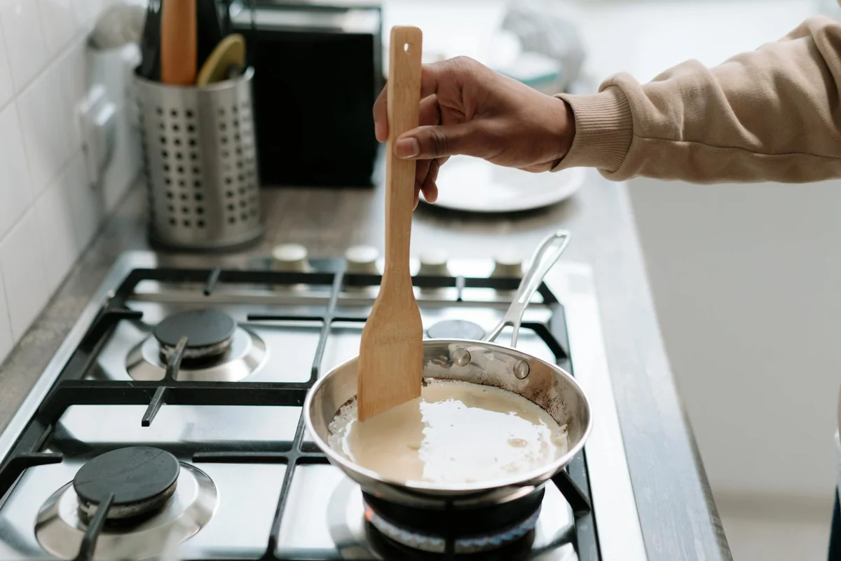 Person stirring cream sauce in a pan on a gas stove, using a wooden spatula.