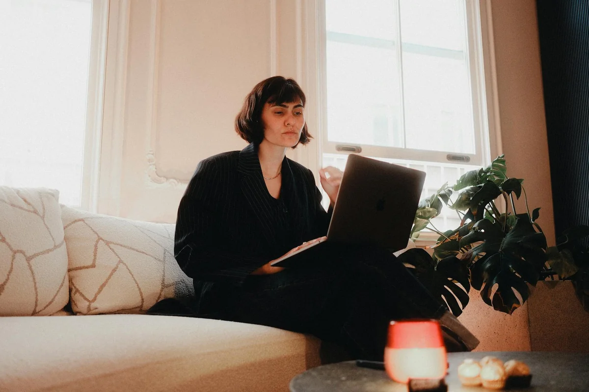 Woman in suit using laptop on sofa in bright room, modern lifestyle.