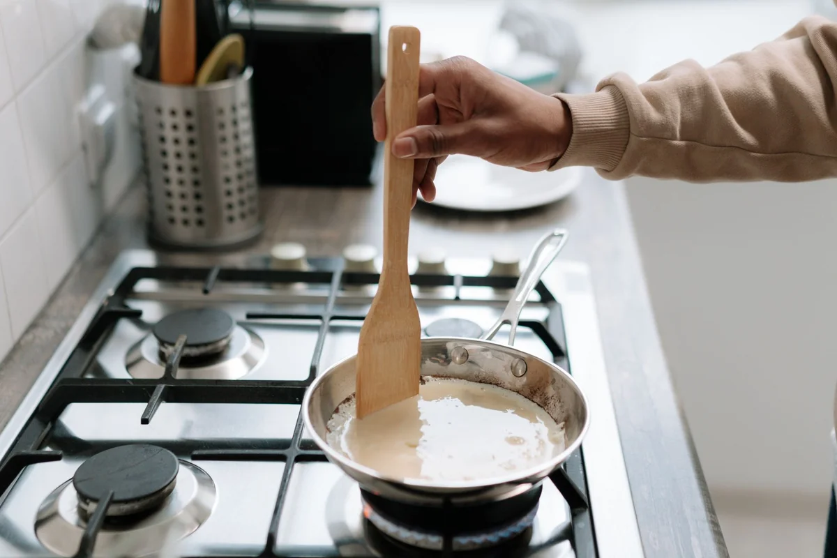 Person stirring cream sauce in a pan on a gas stove, using a wooden spatula.