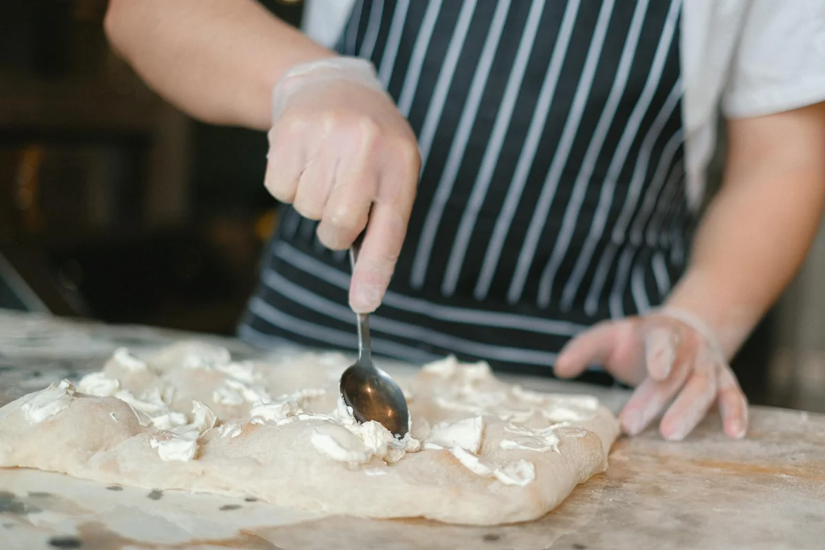 Chef shaping pizza dough with a spoon in a kitchen, spreading creamy cheese. Ideal for culinary themes.
