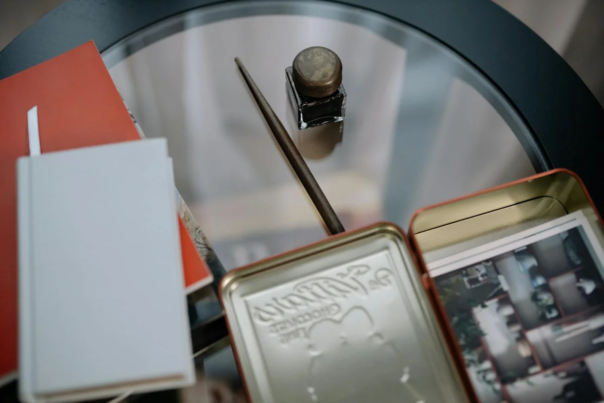 A close-up view of a vintage desk setup featuring notebooks, an ink bottle, and photos on a glass table.