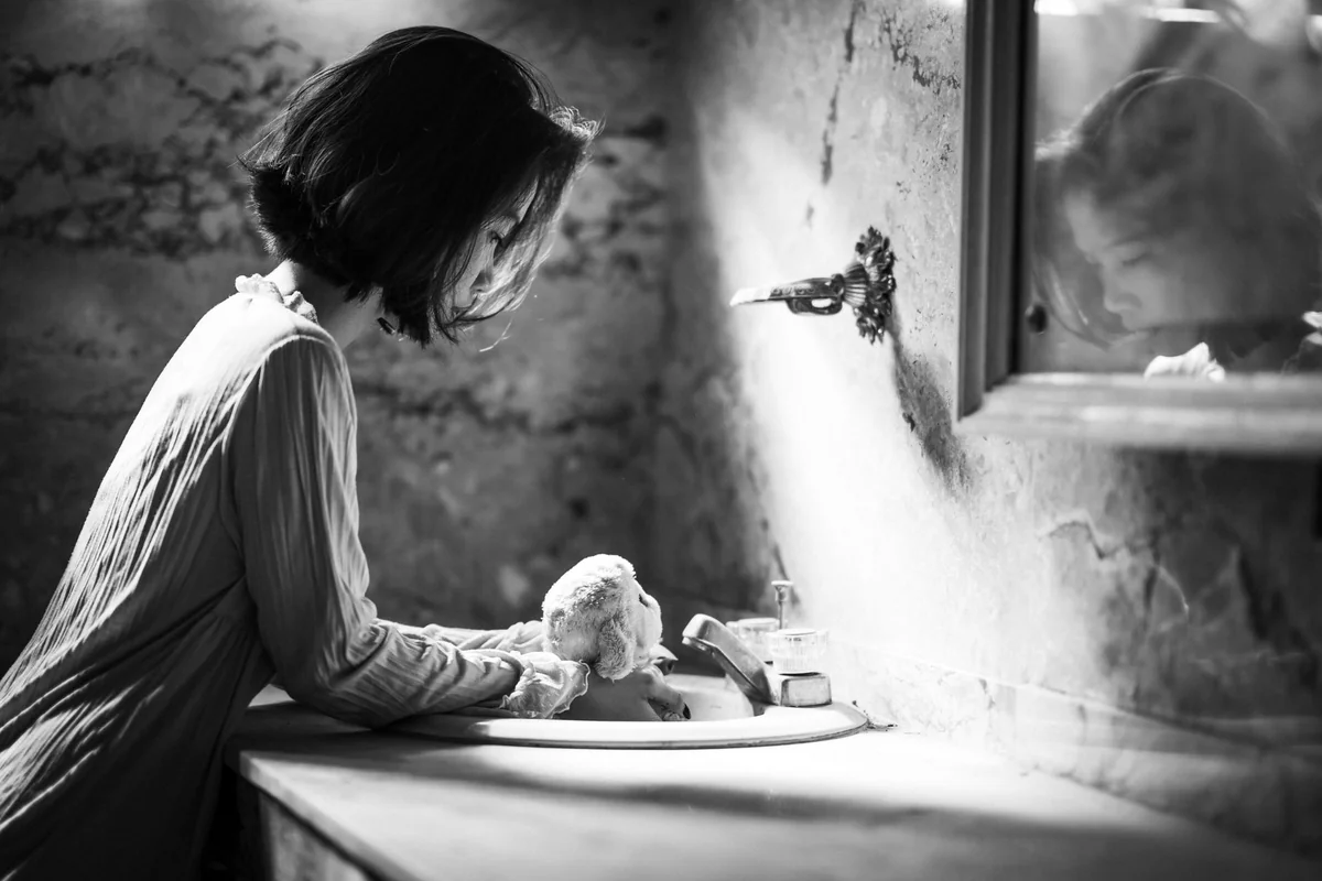 Black and white image of a woman in a vintage bathroom leaning over a sink, reflected in the mirror.