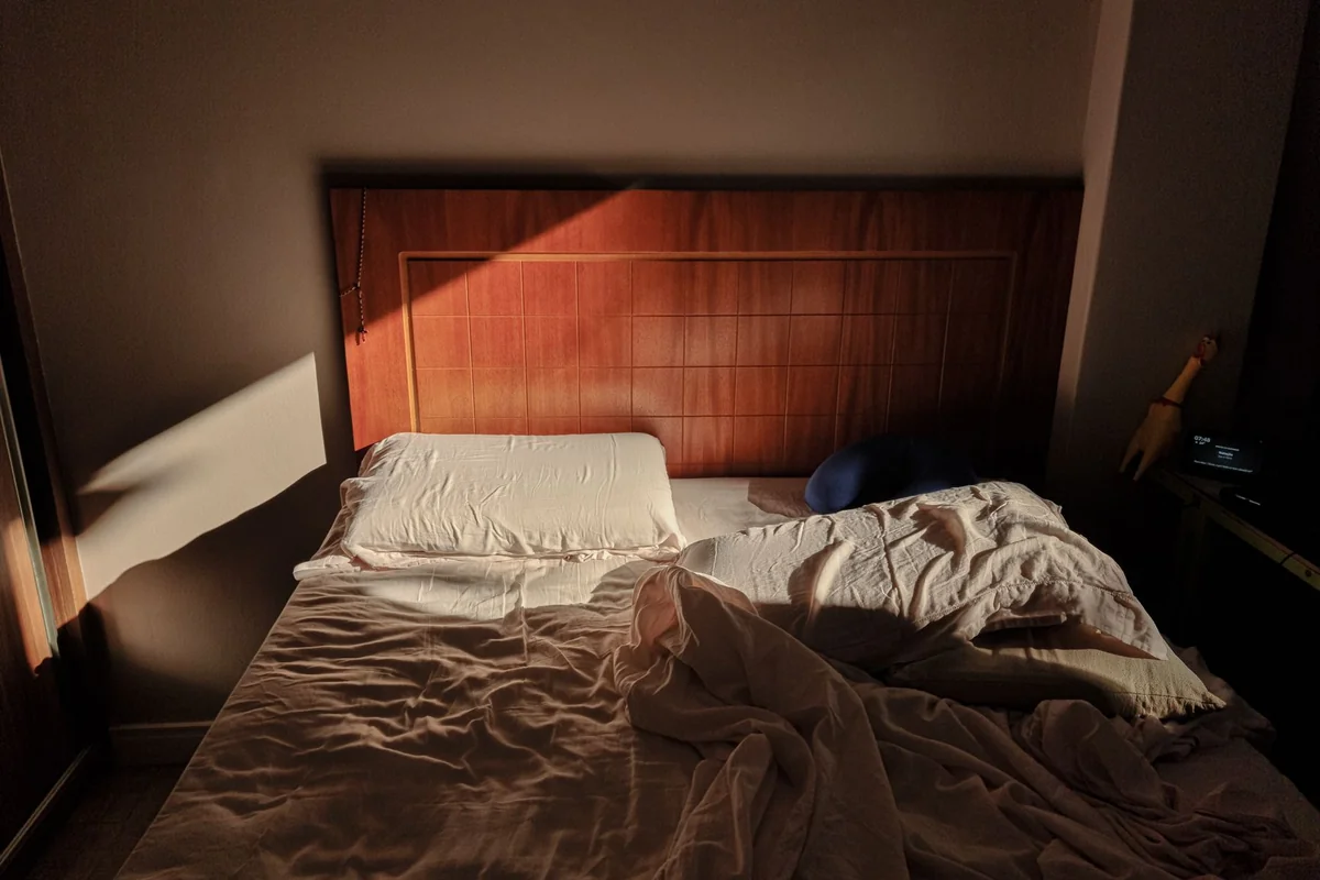 A sunlit, unmade bed with a wooden headboard creates a cozy and warm ambience.