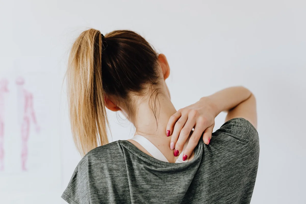 Back view of slim blond female in casual wear with manicure indicating pain spot on back while sitting in front of white wall
