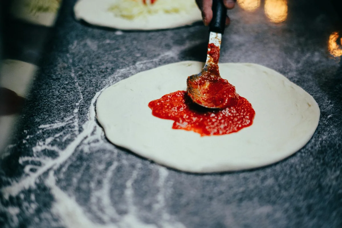 A close-up of a spoon spreading tomato sauce on pizza dough, capturing the pizza-making process.