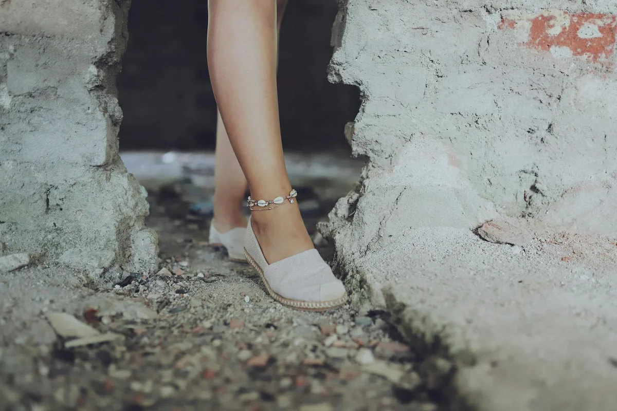 Close-up of a woman's leg wearing an ankle bracelet and espadrilles on a rocky, sandy surface.