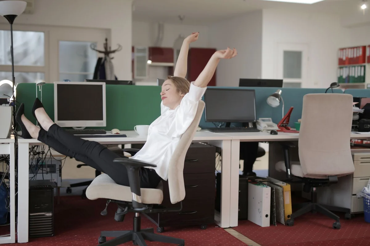 Side view of cheerful female employee in formal outfit sitting on office chair with crossed legs on desk and stretching while
